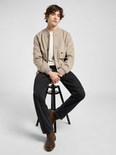 Man sitting on a stool wearing a beige jacket, white shirt, black pants, and brown shoes on a light gray background