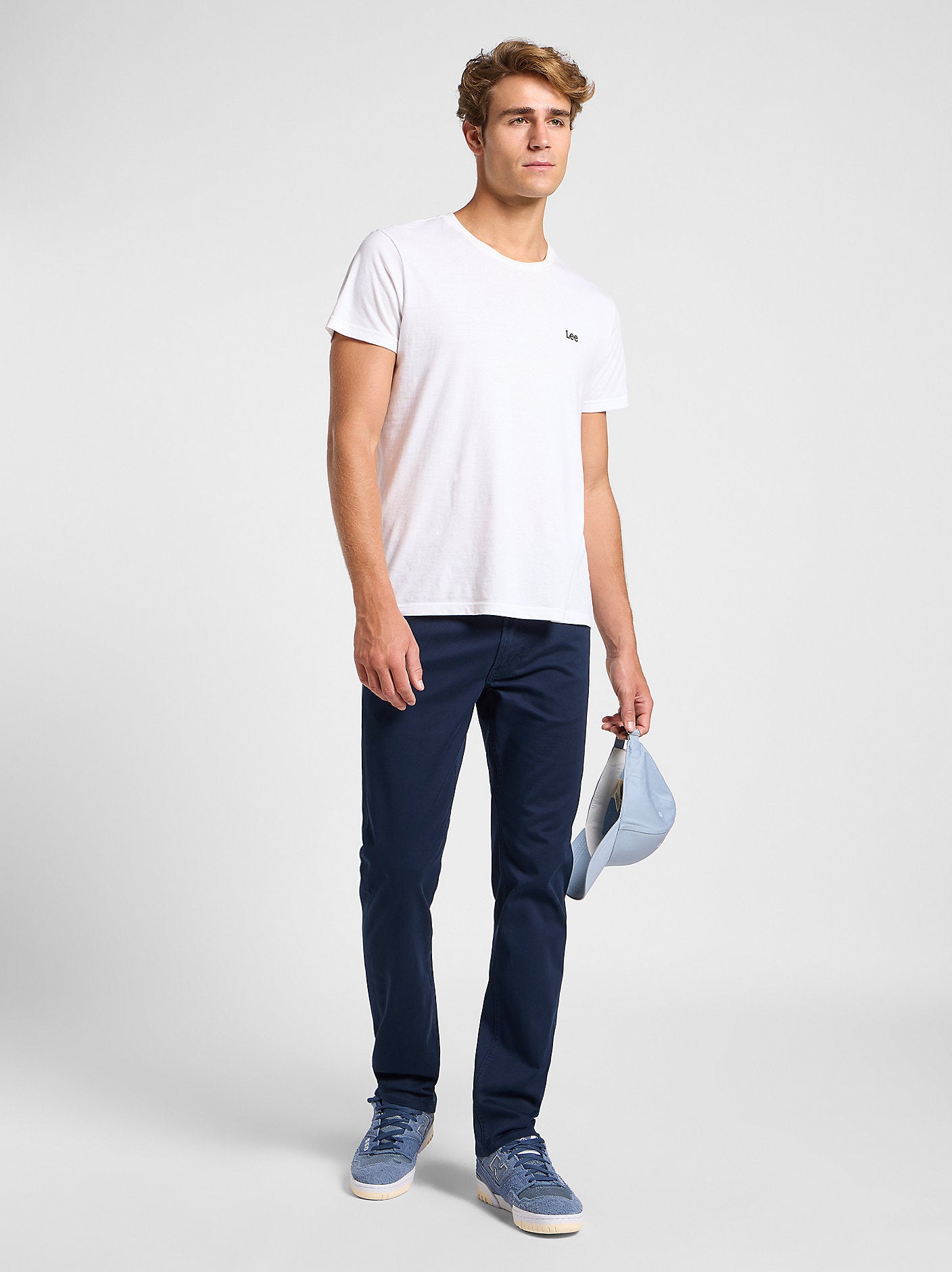 Man wearing a white t-shirt and navy pants holding a cap on a light gray background