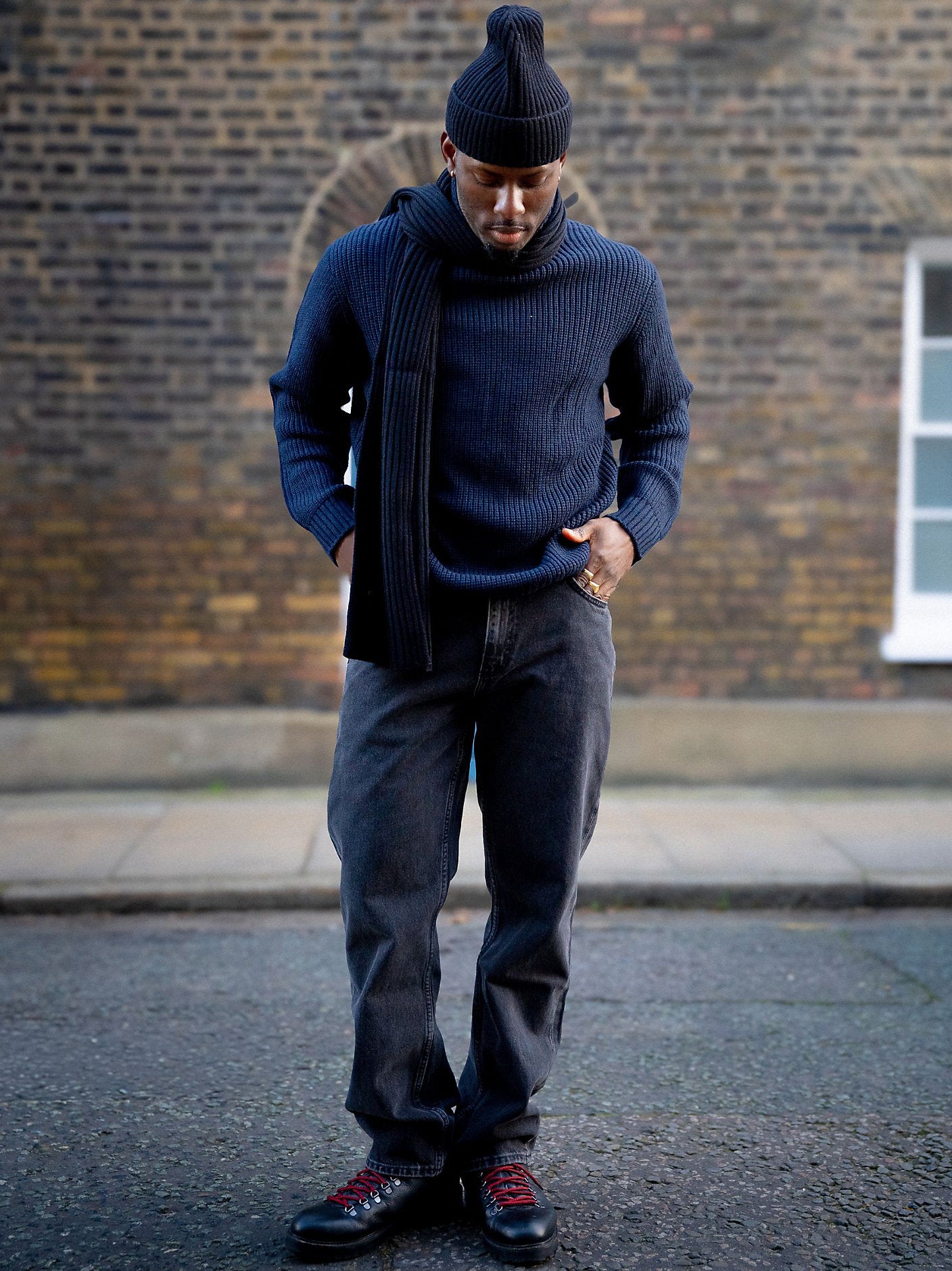 Man wearing a navy sweater, scarf, and beanie standing against a brick wall.
