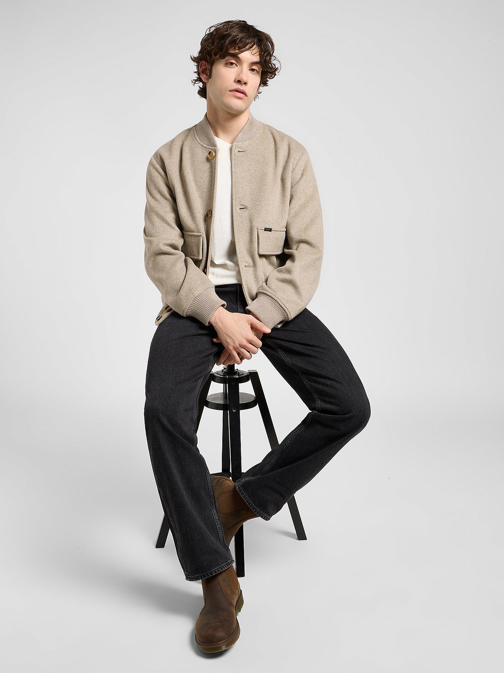 Man sitting on a stool wearing a beige jacket, white shirt, black pants, and brown shoes on a light gray background