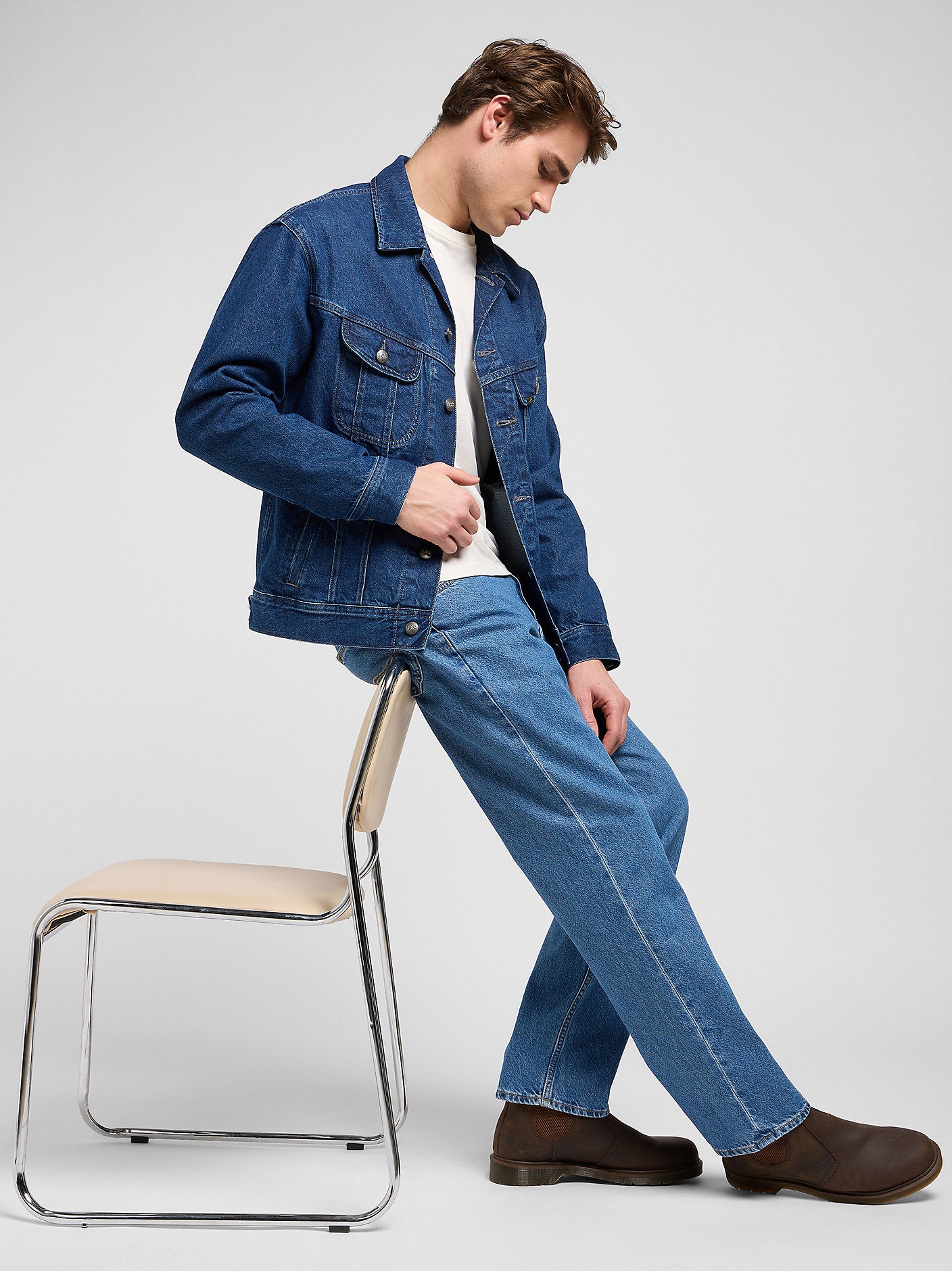 Person wearing a blue denim jacket and jeans sitting on a chair against a white background